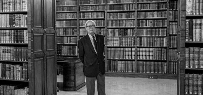 Félix de Azúa de pie en una biblioteca con estanterías de libros