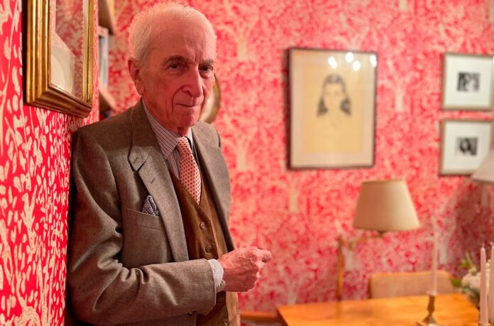 Gay Talese con traje gris junto a una pared roja estampada