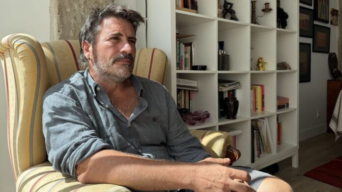 Jacobo García sentado en un sillón, junto a una estantería de libros